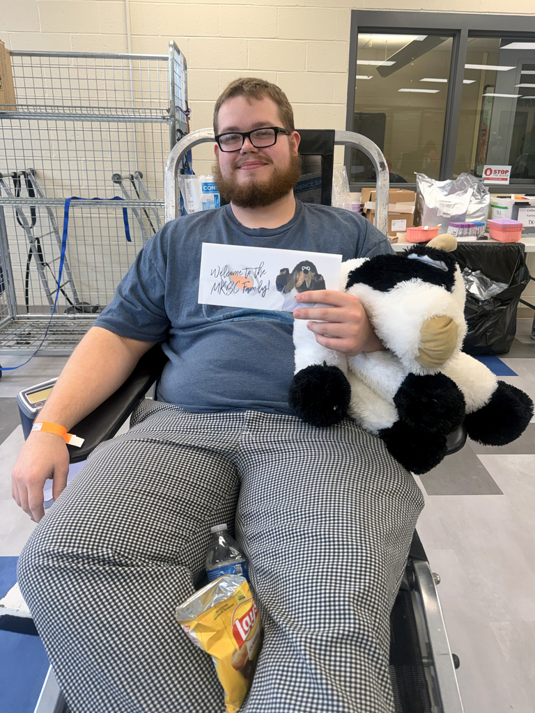 Student donor relaxes in a donation chair holding a plush Highland cow while giving blood at the St. Patrick’s Day themed blood drive.