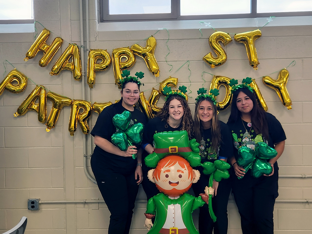 Four BCTC Health Occupations students wearing shamrock headbands stand in front of gold “Happy St. Patrick’s Day” balloons and hold green clover decorations during a themed blood drive.