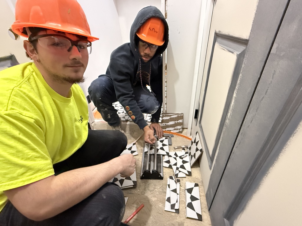 Two Masonry students in hard hats measure and cut patterned tiles for a bathroom floor installation using a tile cutter.