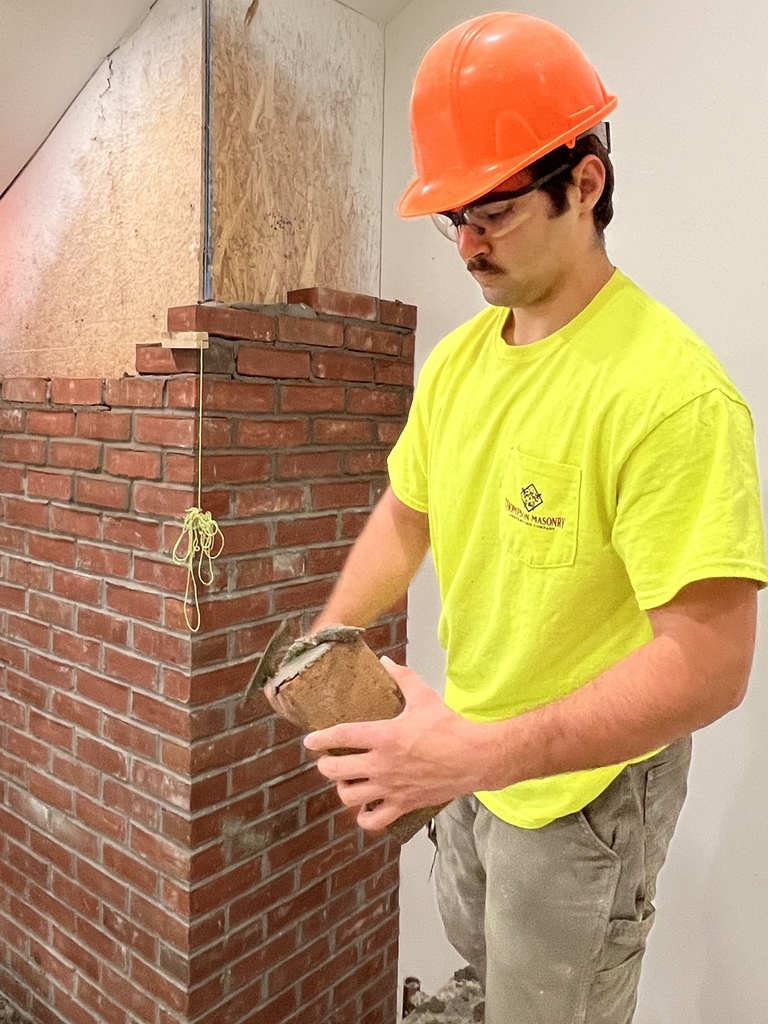 Student in safety glasses and an orange hard hat applies mortar while laying brick on a fireplace structure inside the Student Built House project.