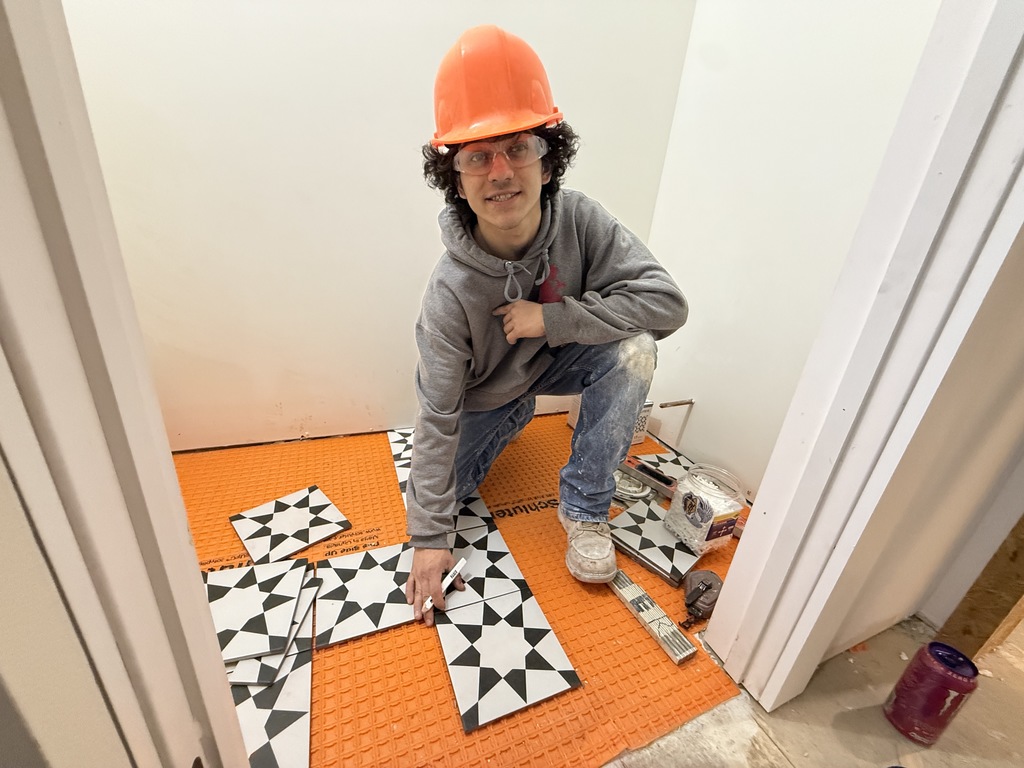 Masonry student wearing a hard hat places patterned tiles onto orange underlayment while preparing the bathroom floor layout.