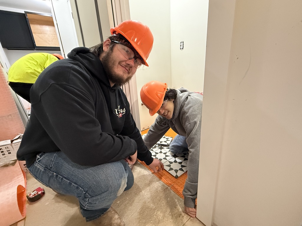 Two Masonry students wearing orange hard hats kneel in a bathroom space while carefully positioning patterned floor tiles on orange underlayment.