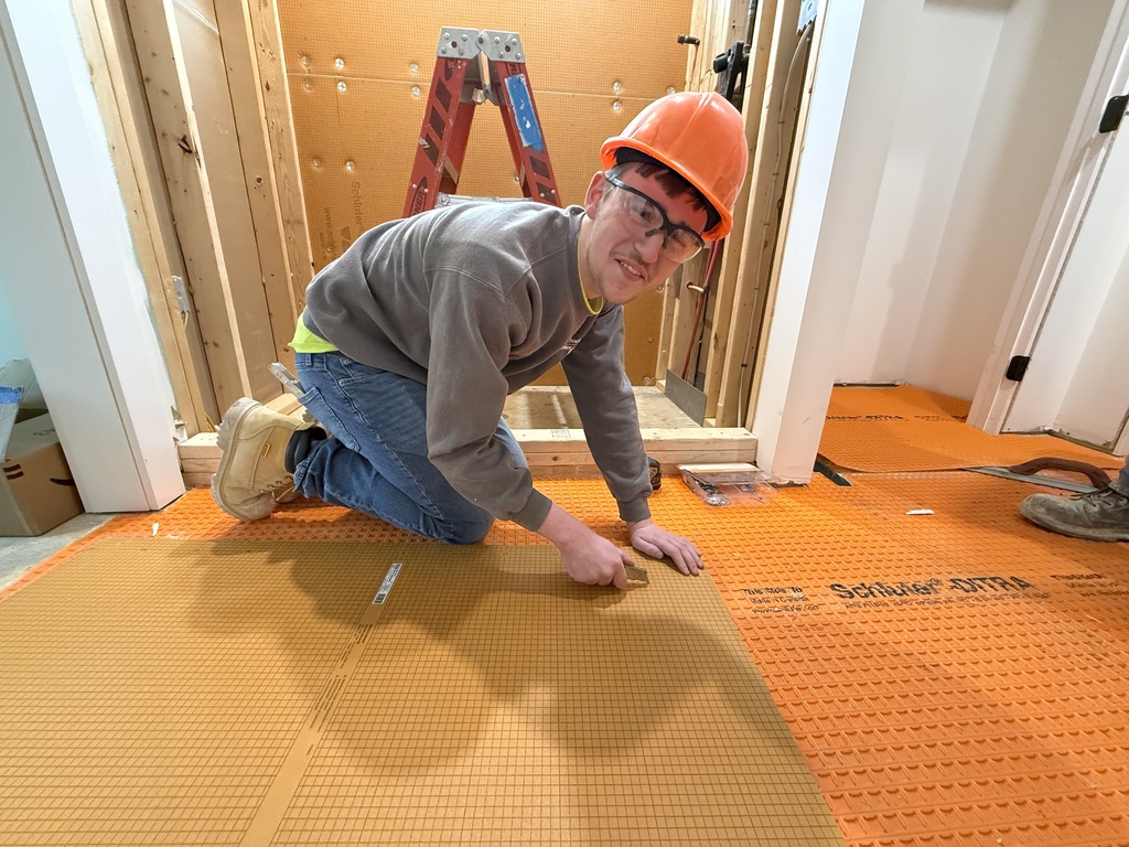 Student wearing a hard hat kneels on orange tile underlayment and smooths adhesive with a trowel while preparing the floor for tile installation.