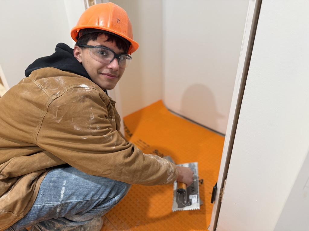 Masonry student wearing a hard hat spreads mortar across orange floor underlayment with a trowel while preparing for tile installation.