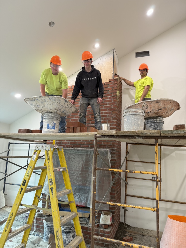 Three Masonry students wearing hard hats stand on scaffolding while building a brick fireplace during the Student Built House project.