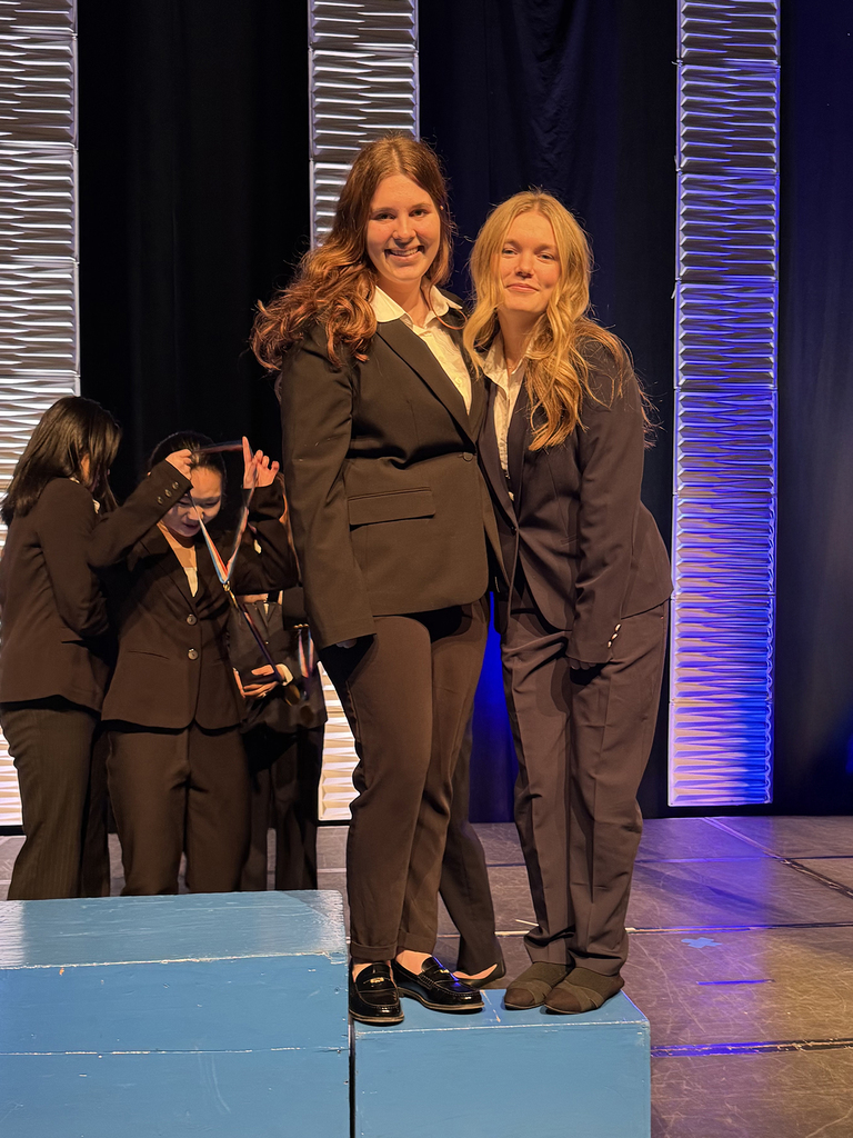 Two students in professional attire stand on a podium on stage while medals are awarded at the Penn HOSA State Leadership Conference.