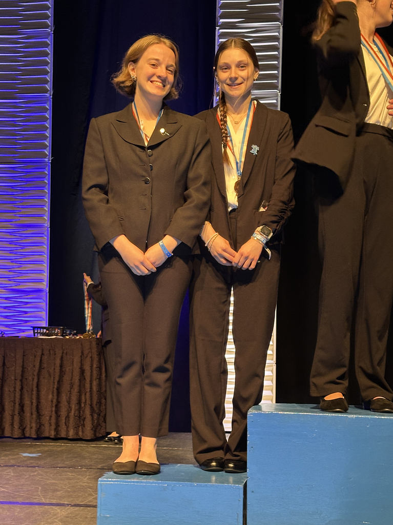 Two students in professional attire stand on a stage podium wearing medals during the Penn HOSA State Leadership Conference awards ceremony.