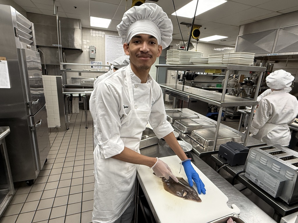 Culinary Arts student in chef uniform smiles while preparing to fillet a flatfish on a cutting board in the BCTC kitchen lab.