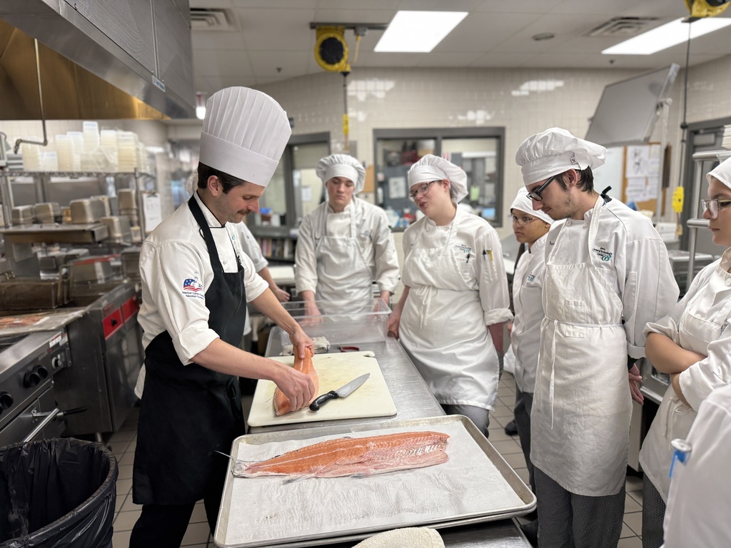 Culinary Arts instructor demonstrates how to fillet a salmon while students in chef uniforms gather around a prep table in the BCTC training kitchen.