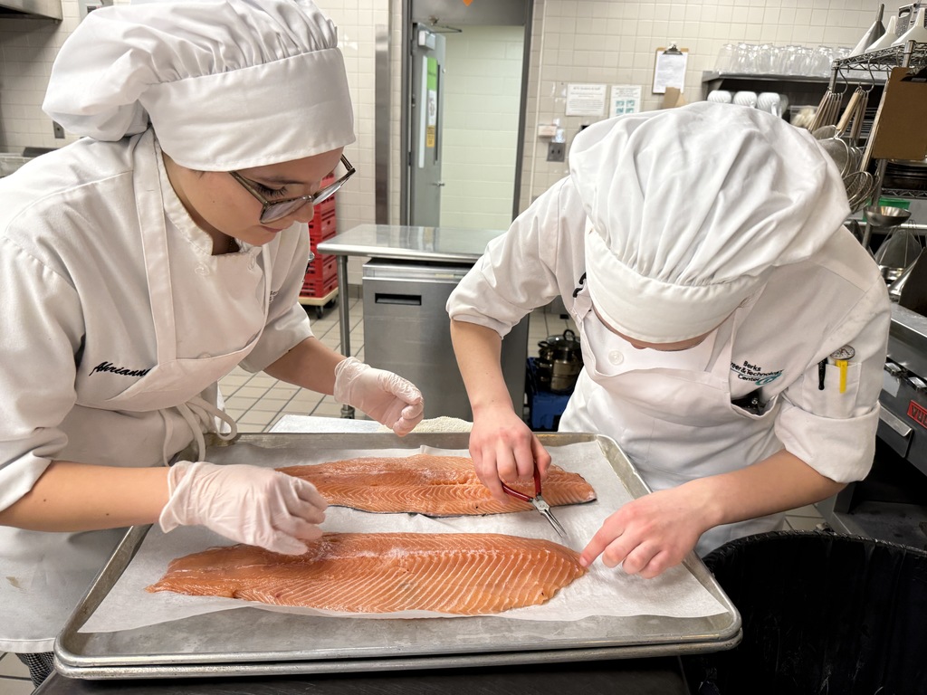 Two Culinary Arts students carefully remove pin bones from salmon fillets using fish tweezers on a tray in the BCTC kitchen lab.