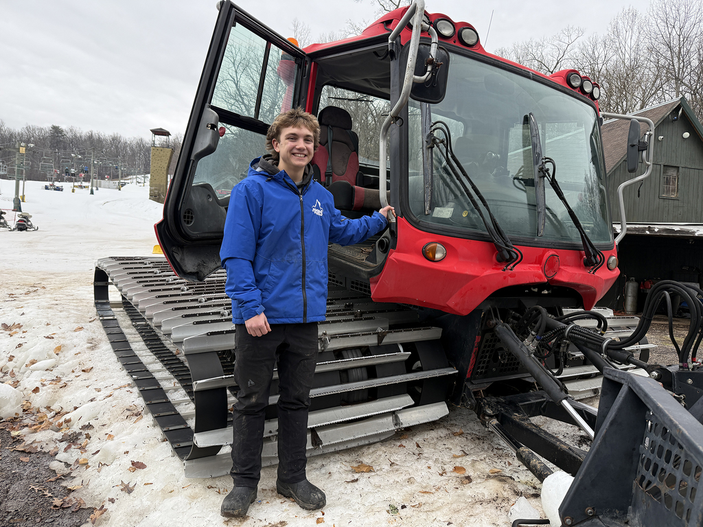 Student stands beside a red snowcat at a ski resort, smiling with the cab door open, showcasing hands-on learning in mountain operations.