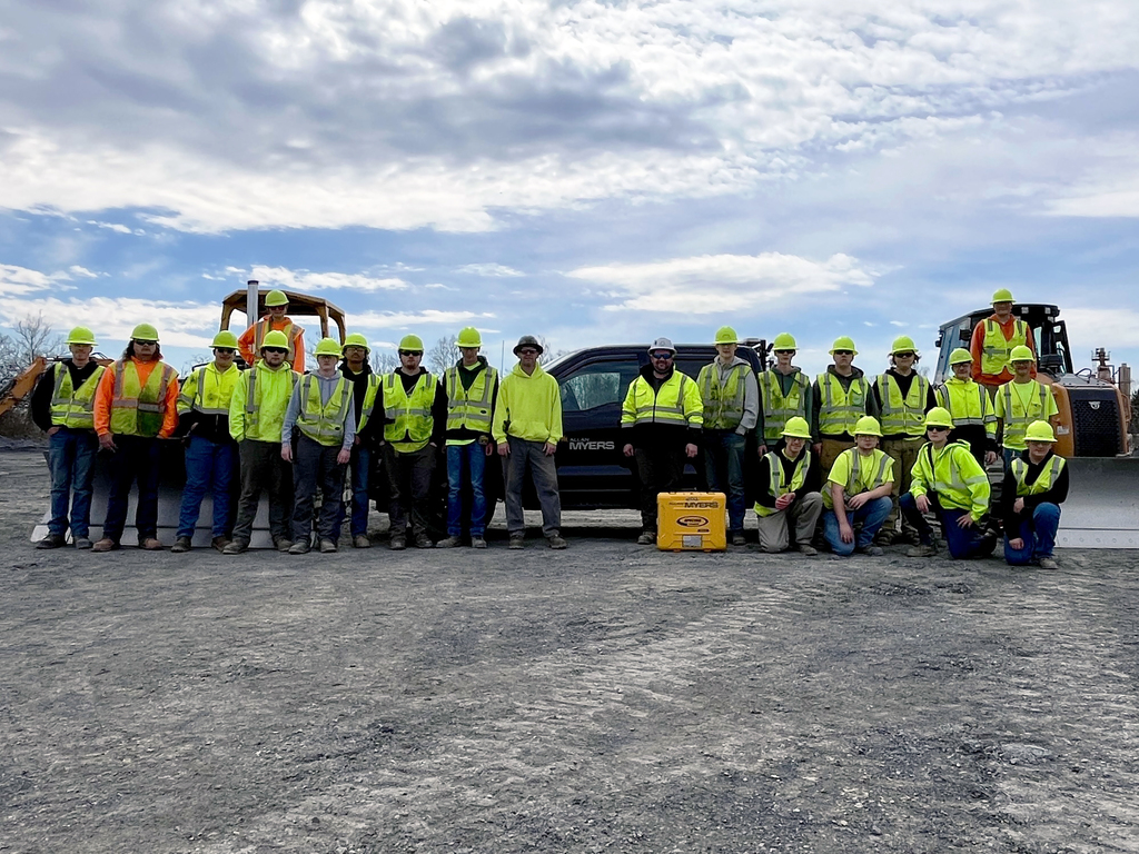 Group of Heavy Equipment Operations students wearing high-visibility vests and hard hats stand at a job site with construction equipment and trucks behind them under a cloudy sky.