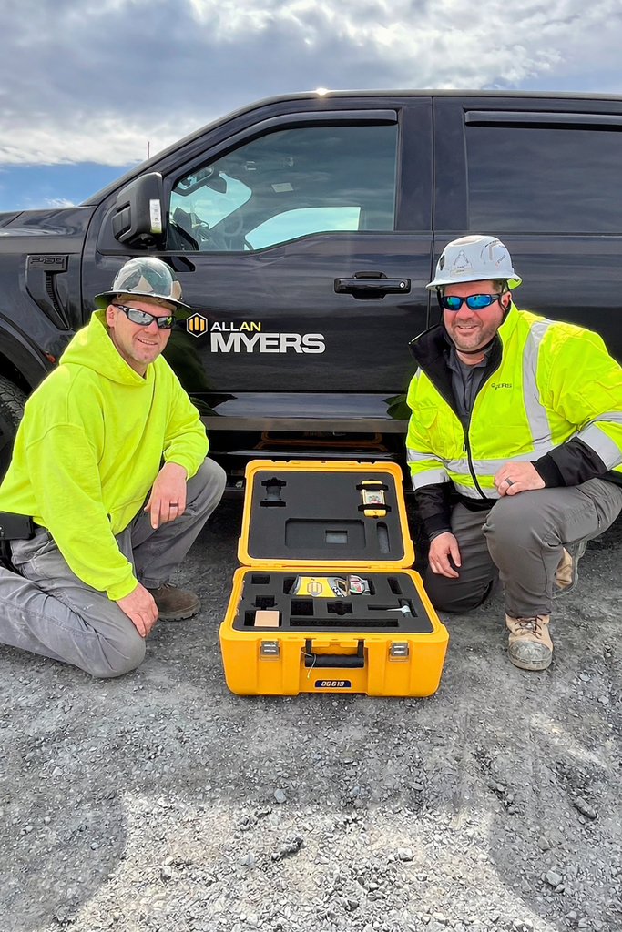 Two men in high-visibility gear and hard hats kneel beside an open yellow case containing a Spectra pipe laser in front of an Allan Myers work truck.