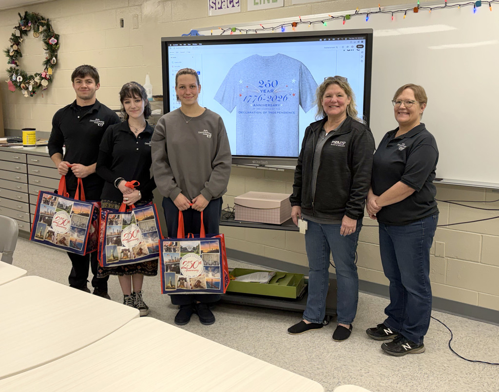 Student Dakota stands closest to a TV screen displaying a t-shirt design, presenting artwork selected for a statewide conference in Gettysburg.