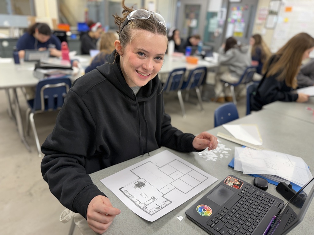 Student smiles at a classroom table while working on a printed kitchen floor plan next to a laptop and design materials.