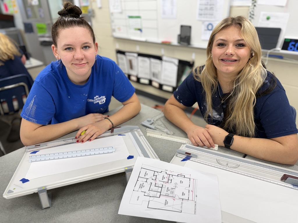 Two students sit at a table with drafting boards and a printed floor plan, smiling toward the camera in a classroom setting.