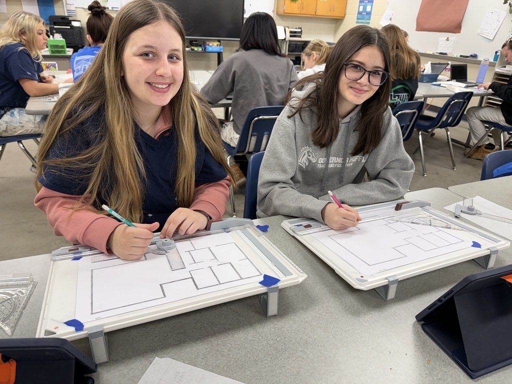 Two students sit at a classroom table using drafting tools to draw floor plan layouts on whiteboards while classmates work in the background.