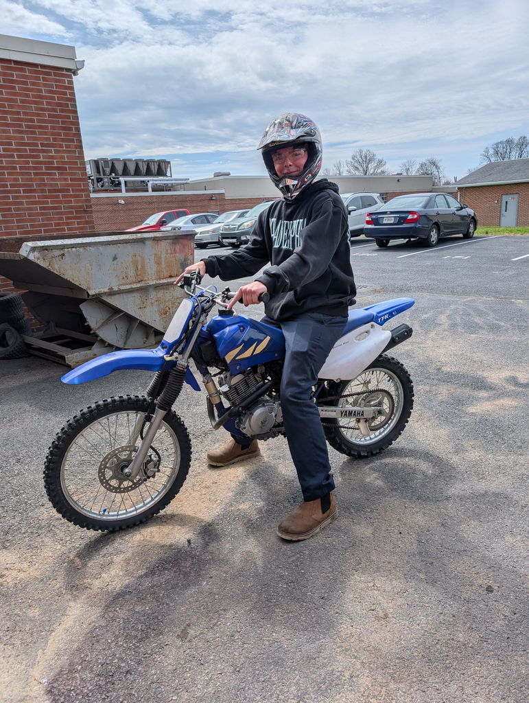 Student sits on a blue dirt bike wearing a helmet, holding the handlebars while parked in a lot near school buildings.