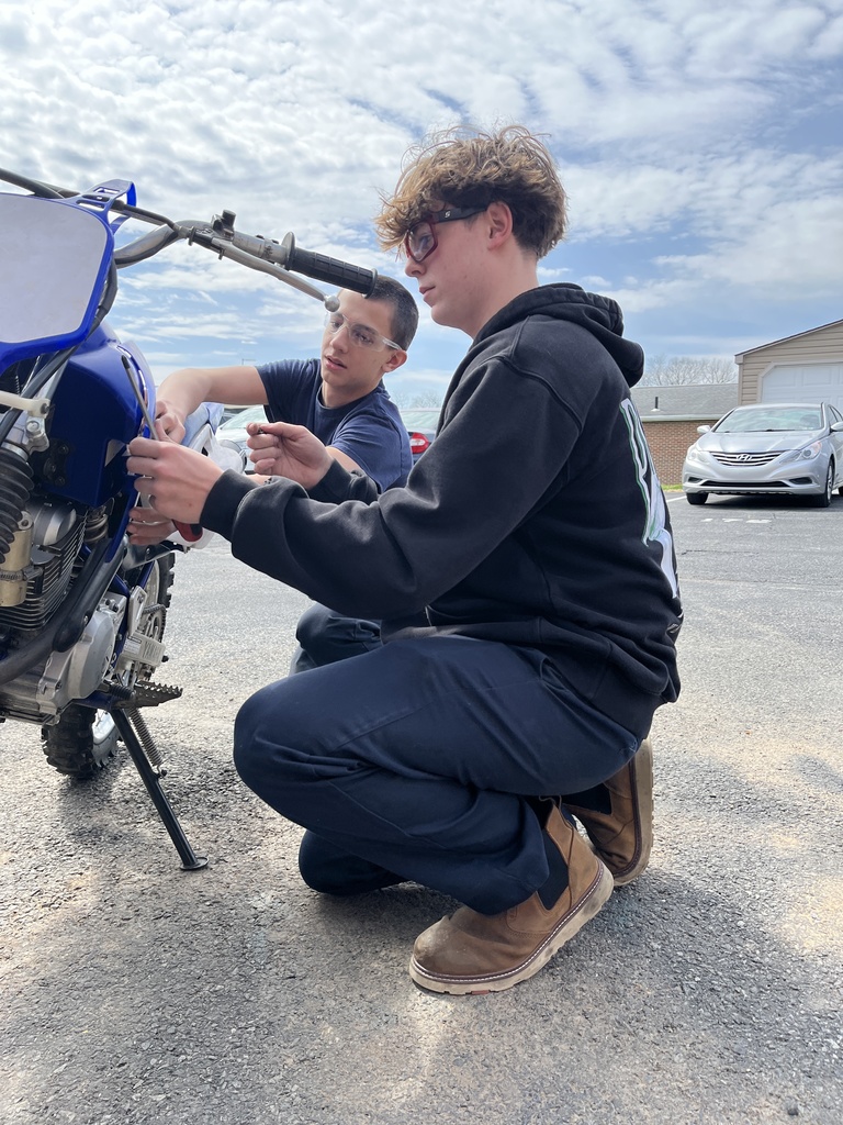 Two students kneel beside a dirt bike, using tools to adjust parts near the front of the engine in a parking lot setting.