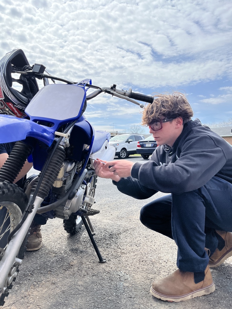 Student crouches next to a blue dirt bike, using a tool to make adjustments near the engine under a cloudy sky.