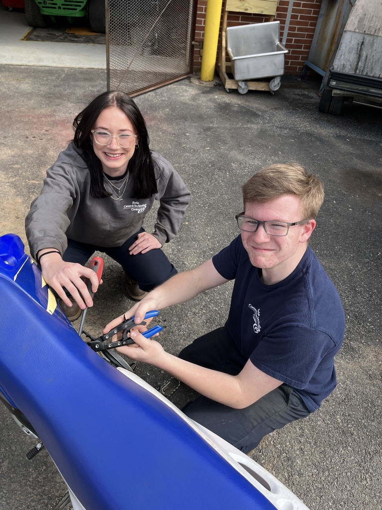 Two students kneel beside a blue dirt bike, using hand tools to adjust a component while looking up toward the camera.