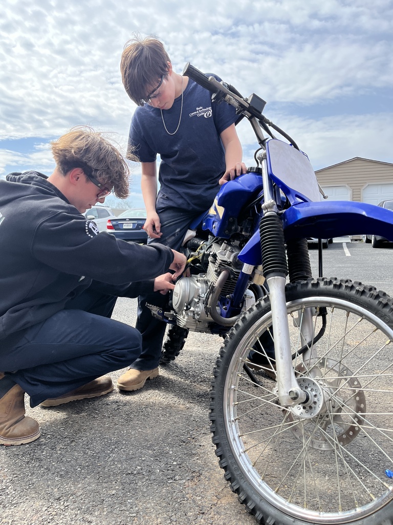 Two students work on the engine of a blue dirt bike outdoors, focusing on wiring and components near the motor.