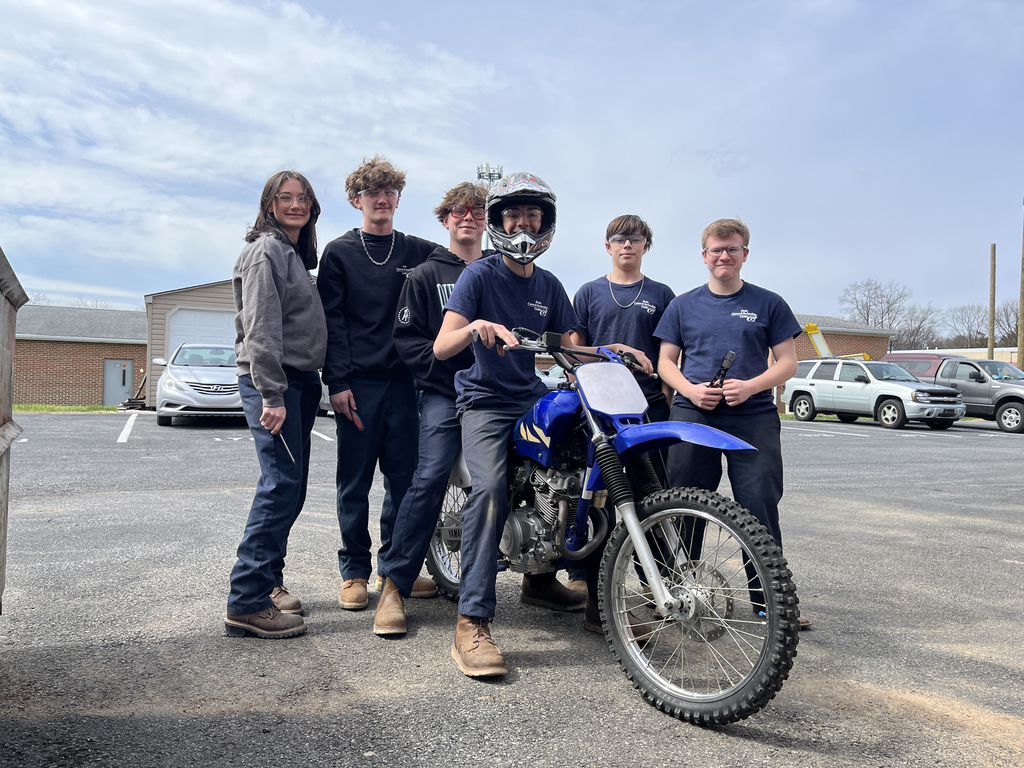 Six students stand around a blue dirt bike in a parking lot; one sits on the bike wearing a helmet while others pose nearby.