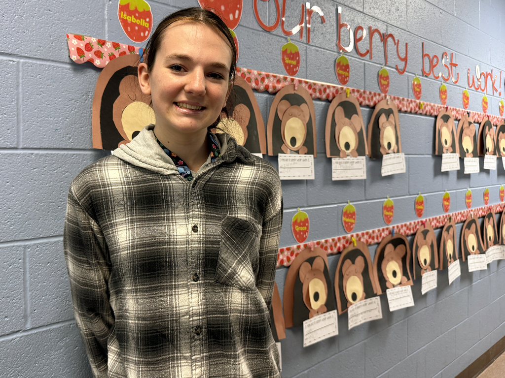 Student smiles while standing beside a classroom bulletin board decorated with strawberry-themed student work and bear face crafts displayed in rows.