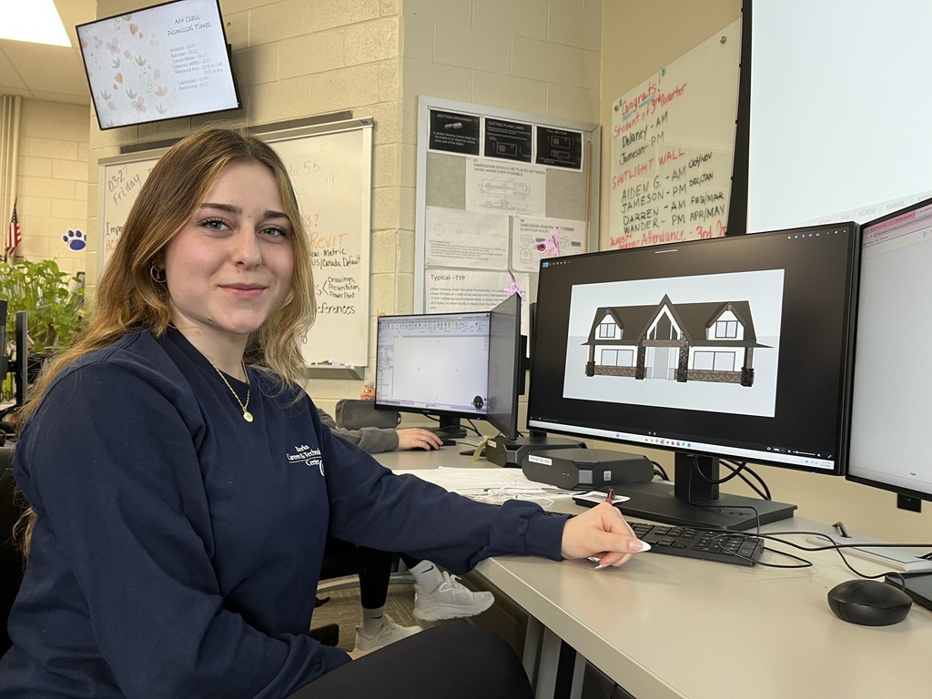 Student seated at workstation displaying a detailed 3D house rendering on a computer screen in drafting class.