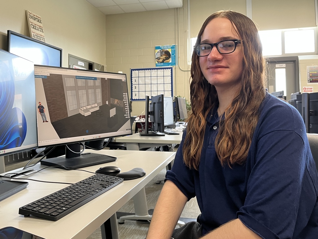 Student at computer viewing a 3D interior design rendering with furniture and lighting elements on screen.