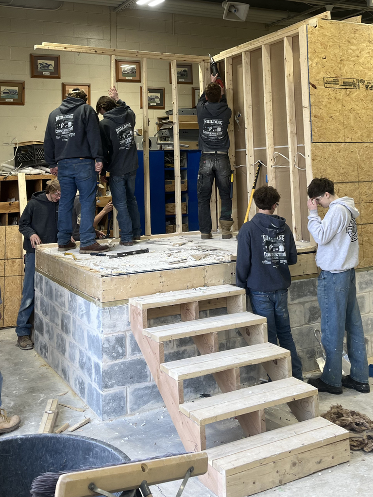 Several students stand on a raised platform removing wall framing from the training structure using hand tools.