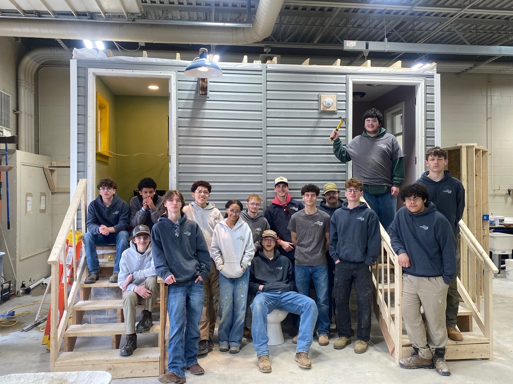 Group of Building Construction Occupations students posing in front of a small framed and sided training structure inside a workshop.