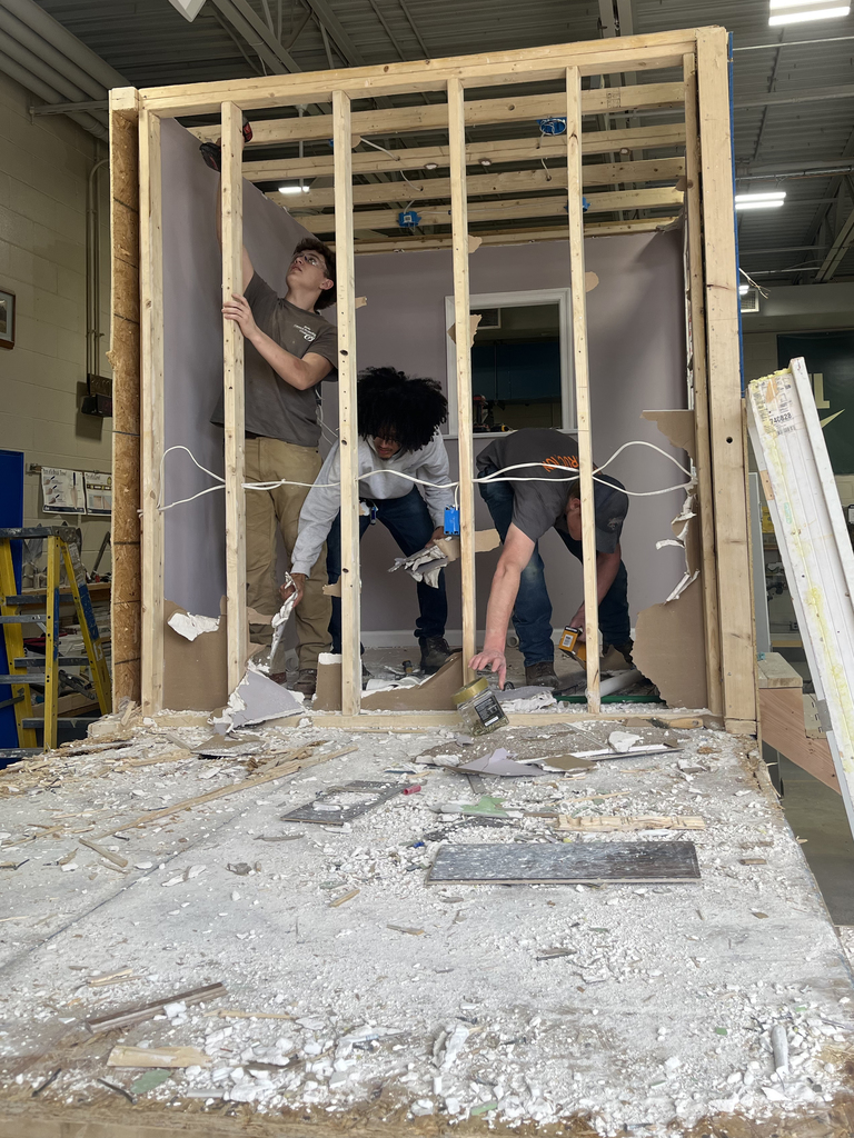 Students inside a framed structure tear down drywall, exposing studs and wiring during a hands-on demolition activity.