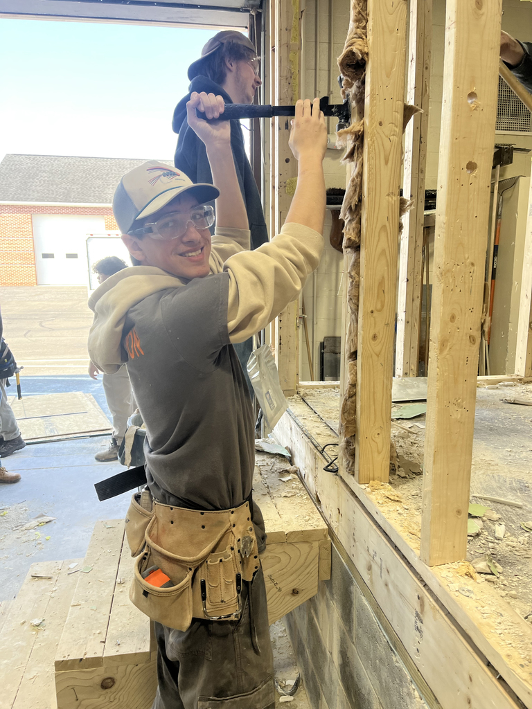 Student wearing safety glasses uses a hammer to remove insulation from a framed wall during demolition inside the shop.