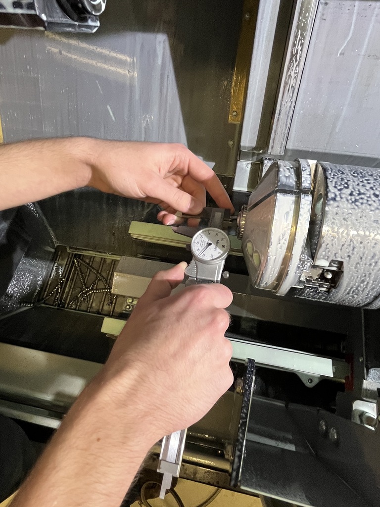 Close-up of a student measuring a machined metal part with a dial caliper inside a CNC machine workspace.