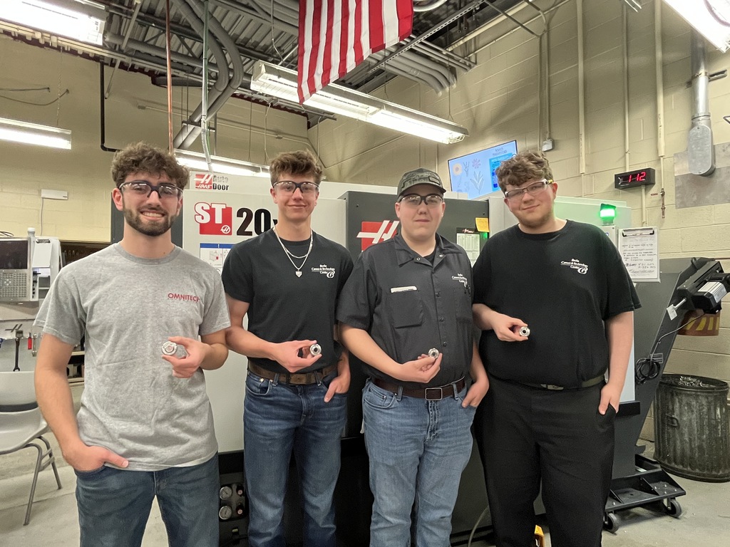 Students in a machining lab hold finished metal parts in front of a CNC machine, wearing safety glasses under an American flag.