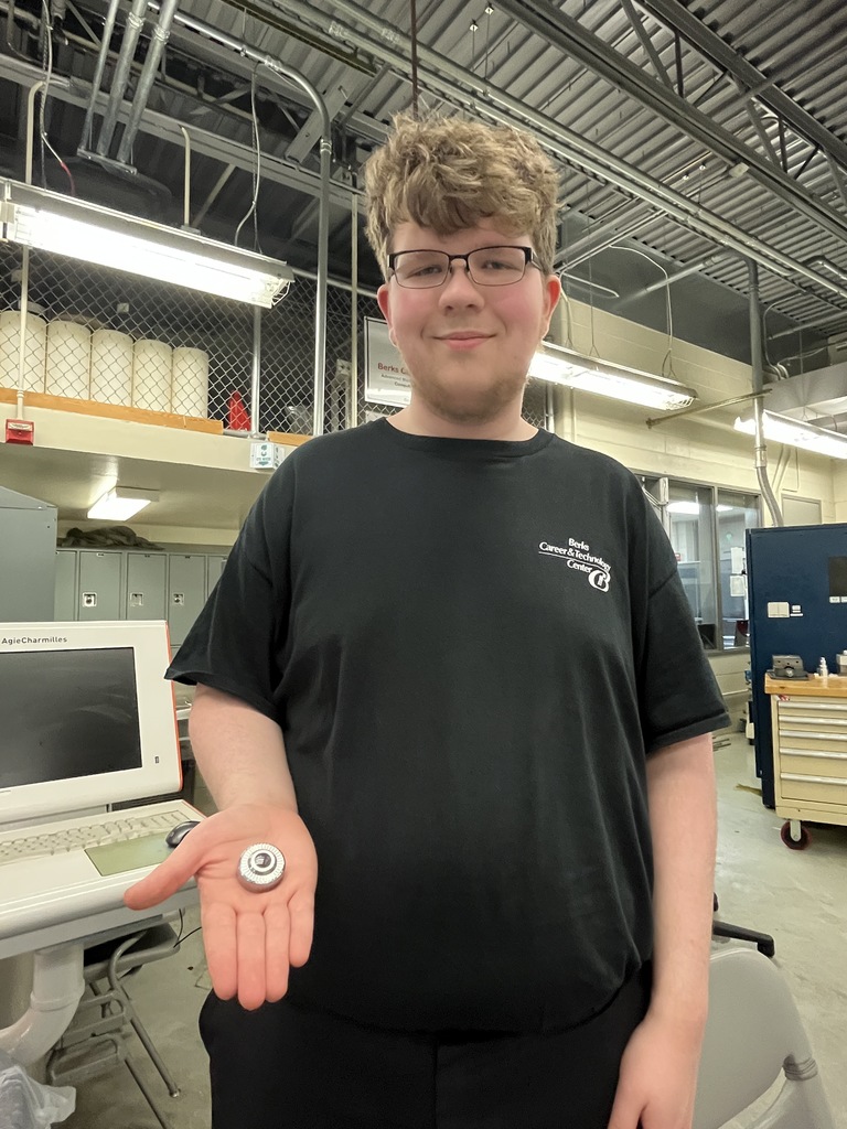 Student stands in a machining lab holding a finished metal part in their hand, with CNC equipment in the background.