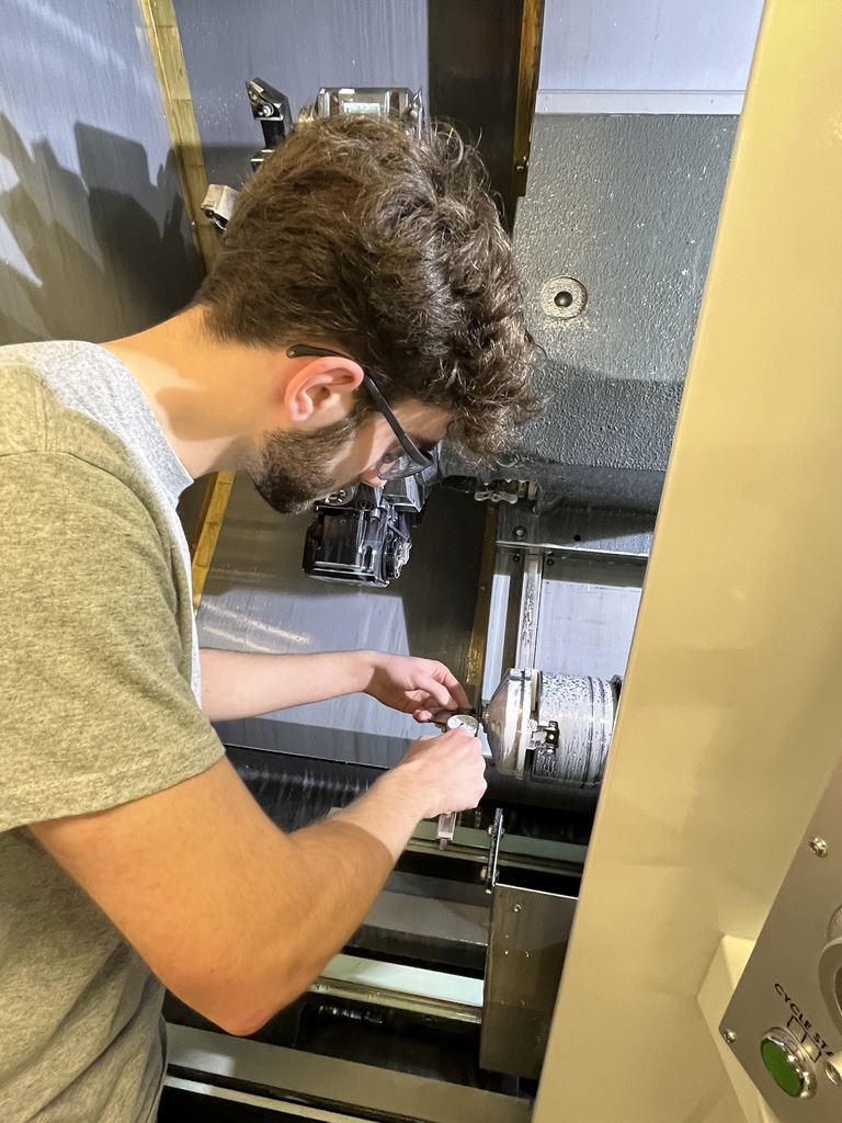 Student uses a precision measuring tool to check a metal part inside a CNC machine, focusing closely on accuracy.
