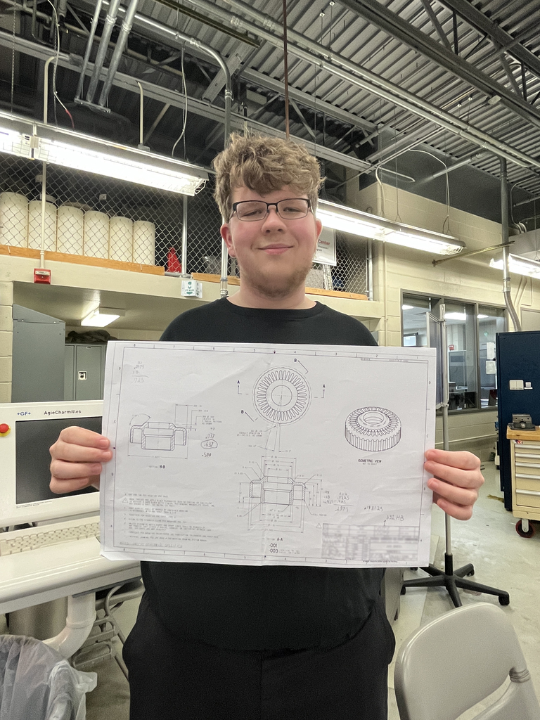 Student holds a technical drawing of a machined part in a lab, with equipment and workstations visible behind.