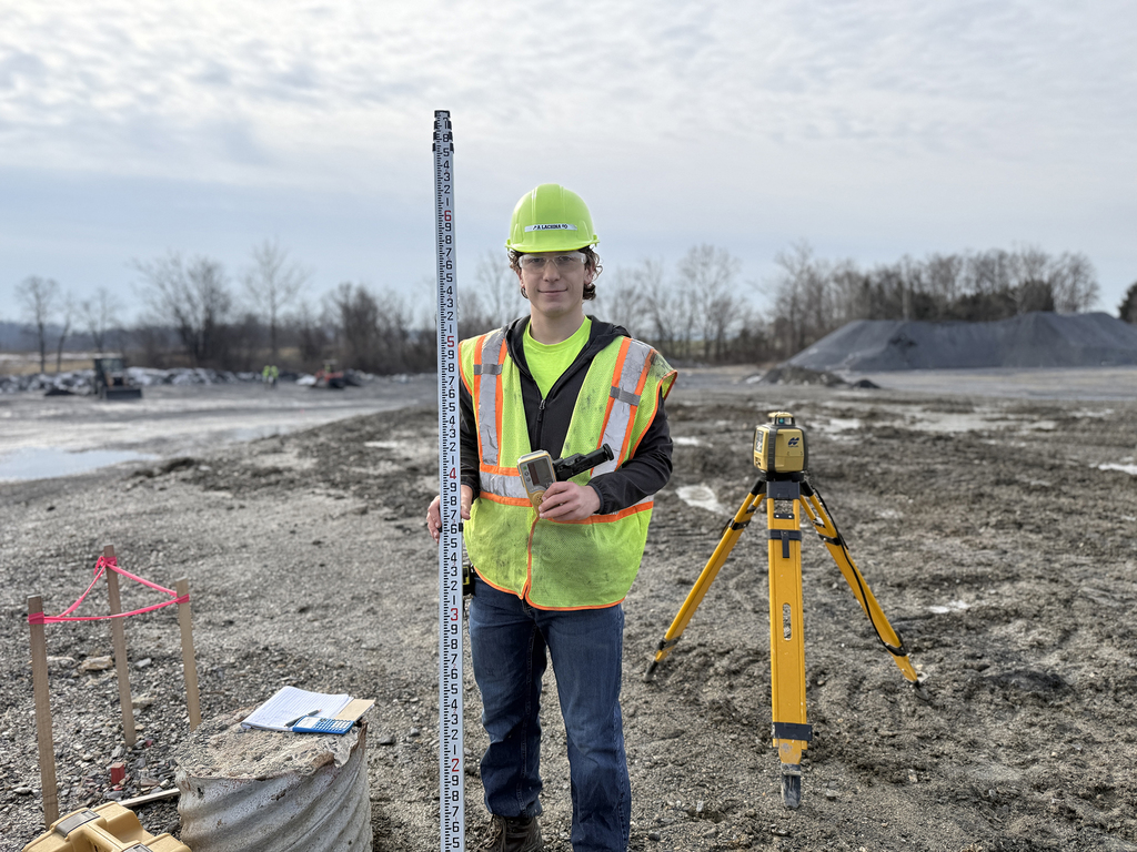 Student in safety vest and hard hat holds a grade rod and survey equipment at a quarry site.