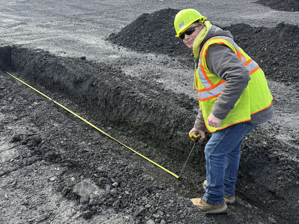Student in safety gear measures the width of a trench using a tape measure in a quarry.