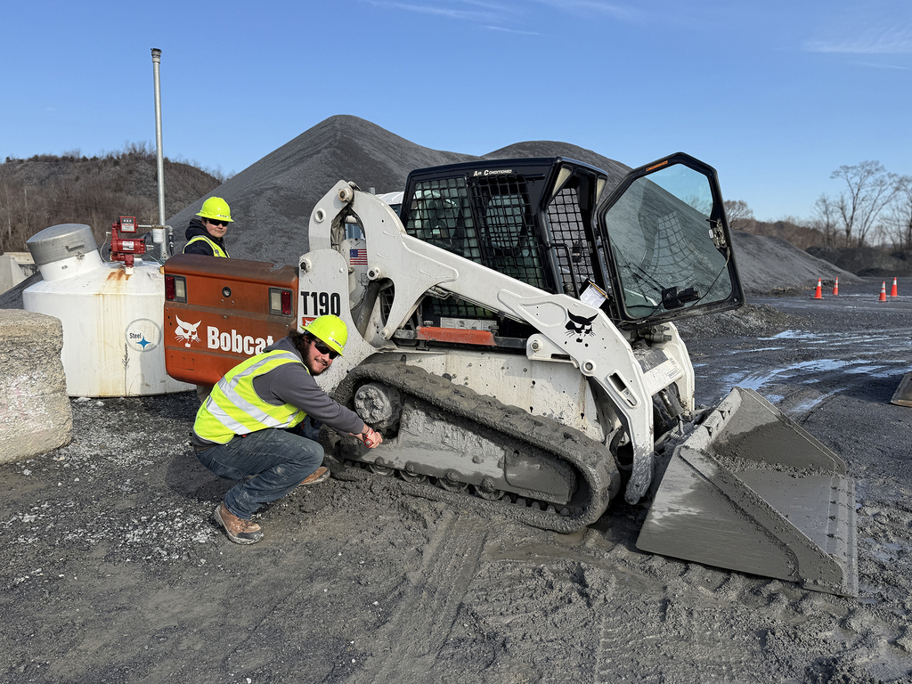 Two students in safety gear work on a skid steer, performing maintenance or inspection at a quarry.