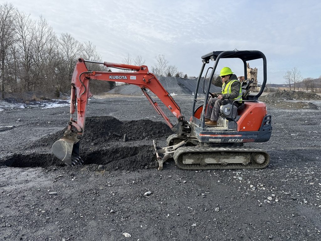 Student operates a compact excavator, digging a trench in a gravel quarry.