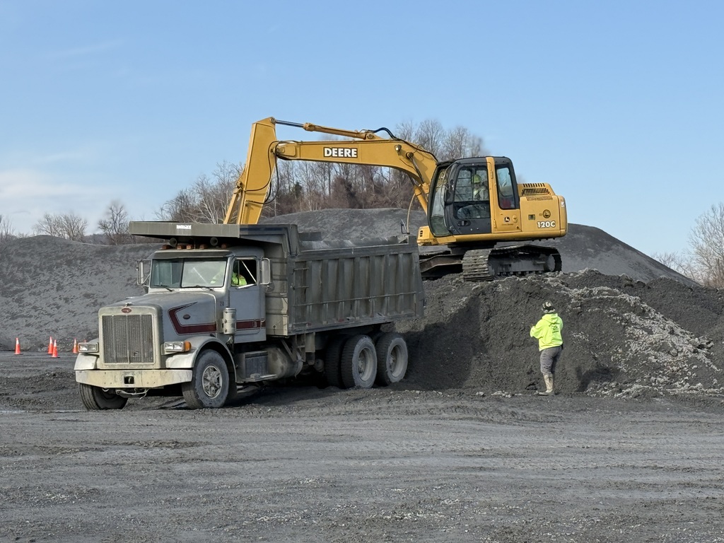 Excavator loads gravel into a dump truck at a quarry while another worker stands nearby.