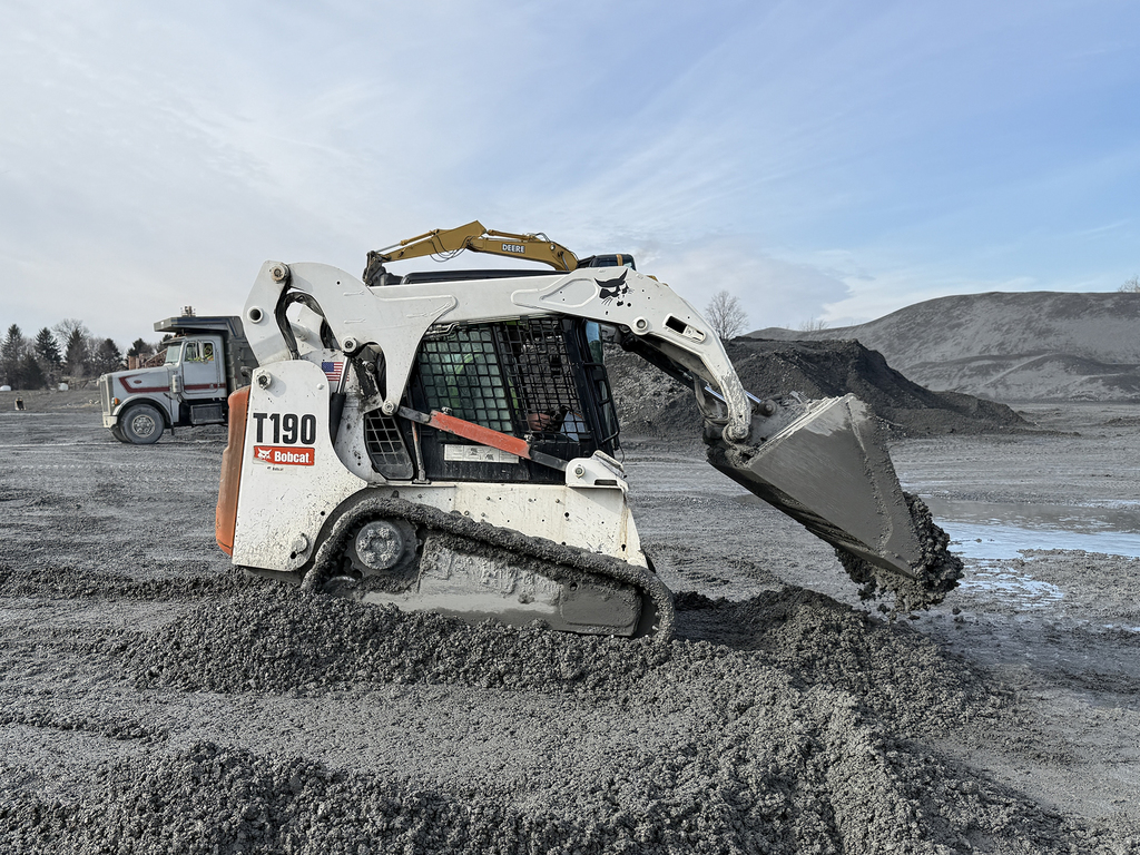 Skid steer loader pushes gravel forward across uneven ground in a quarry.