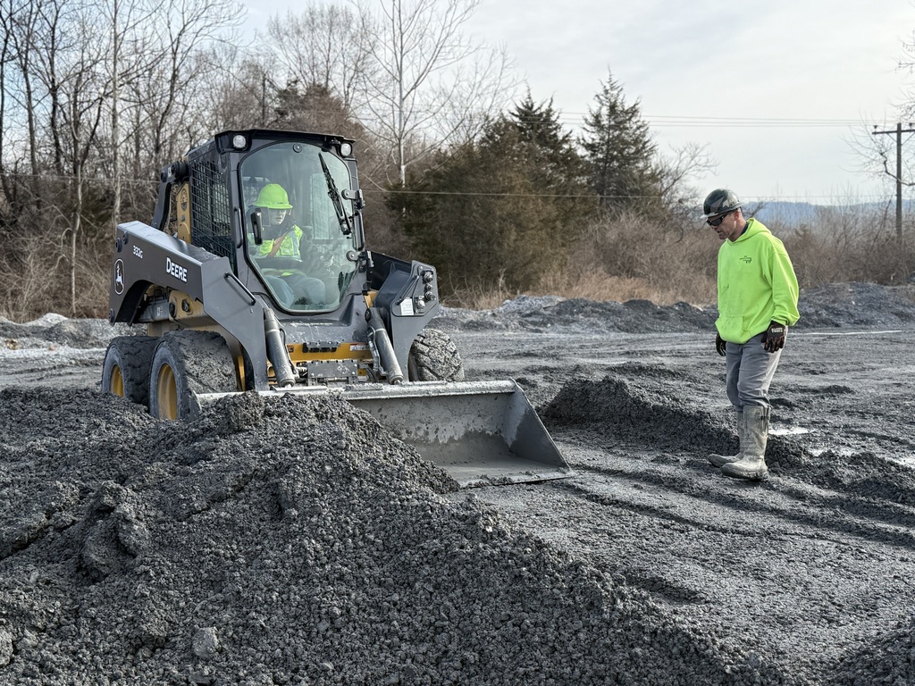 Student operates a skid steer while an instructor stands nearby observing at a quarry site.