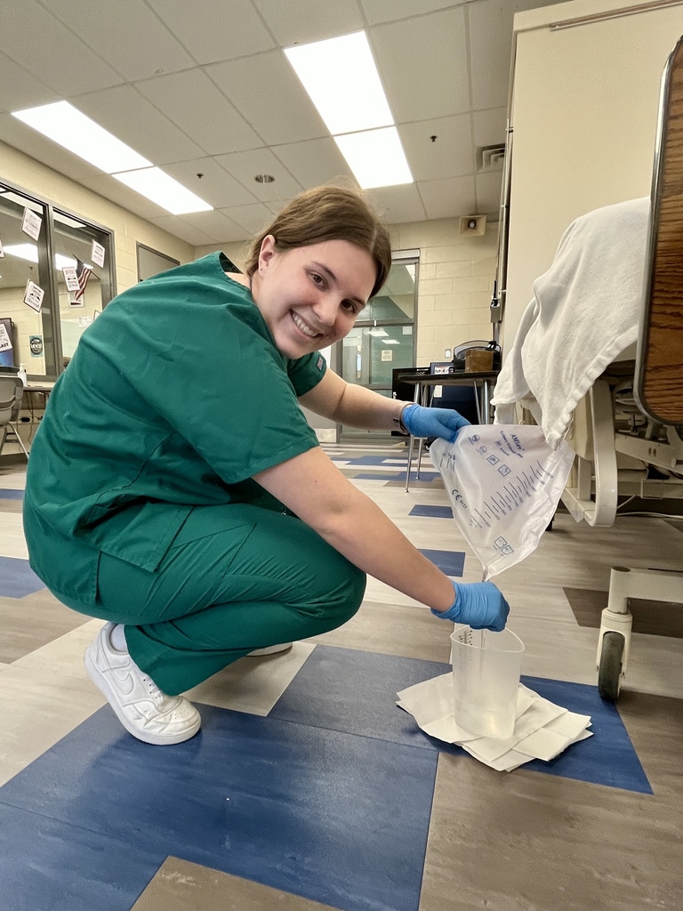 Student in scrubs empties a catheter drainage bag into a container while kneeling beside a hospital bed.
