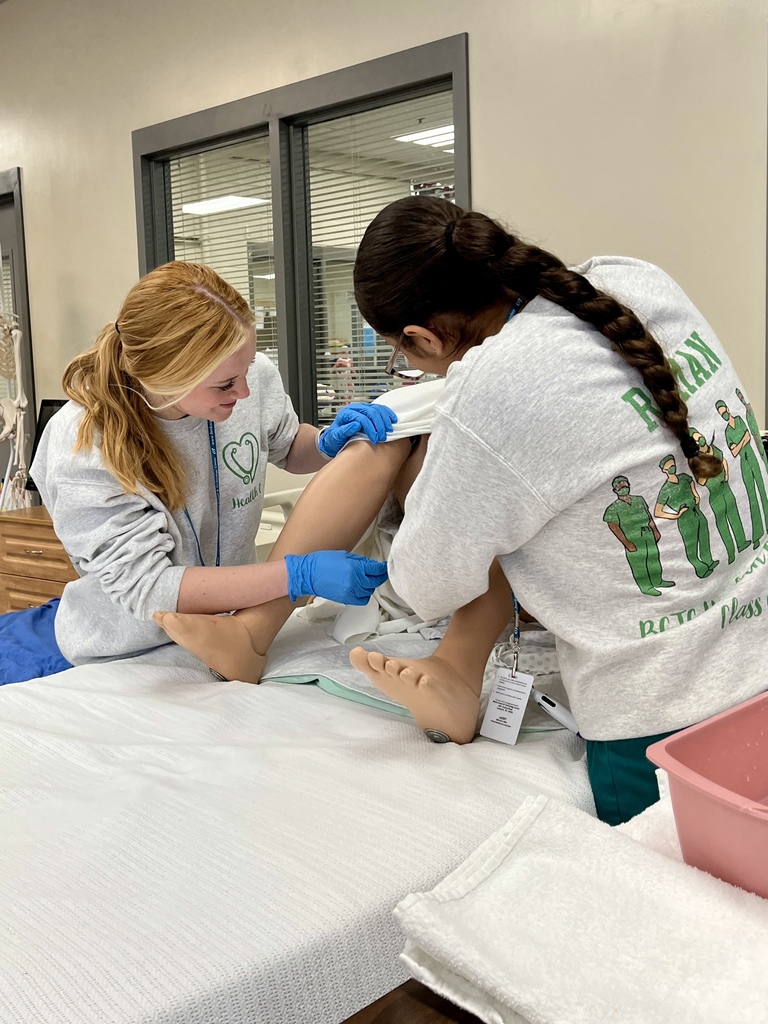 Two students in gloves practice catheter care on a medical mannequin leg in a simulated patient care setting.