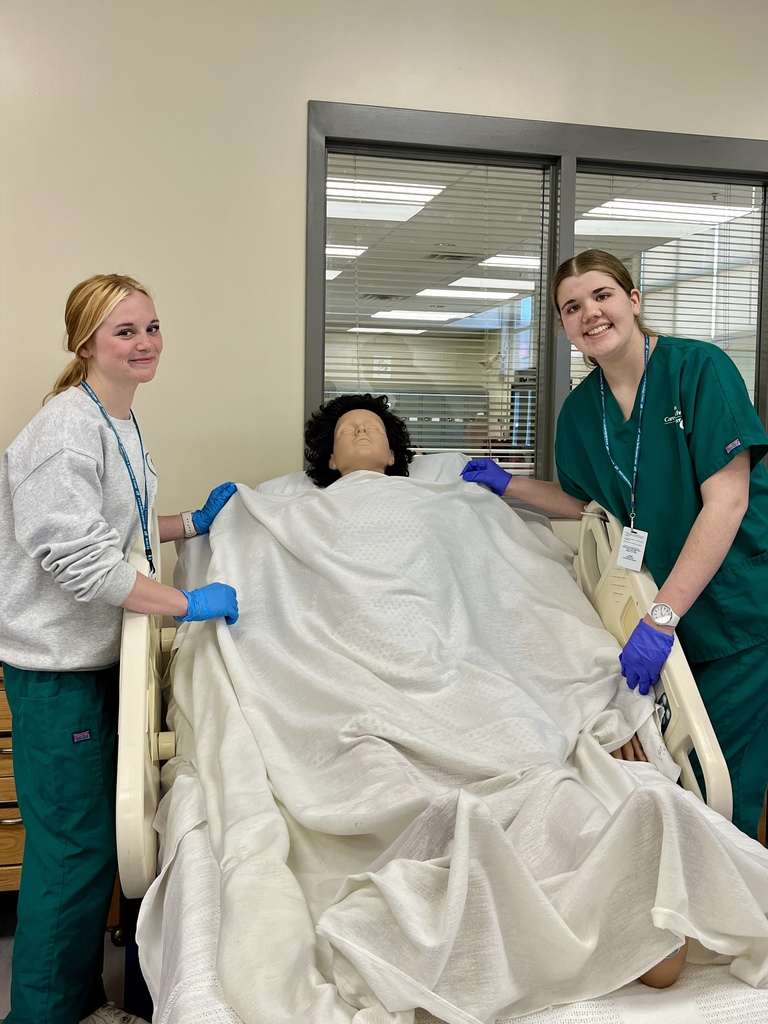 Two students stand beside a mannequin patient in a hospital bed, adjusting linens during a care simulation.