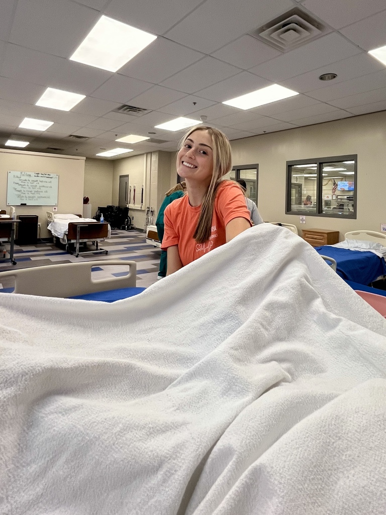 Student smiles while making a hospital bed, lifting sheets in a healthcare classroom.